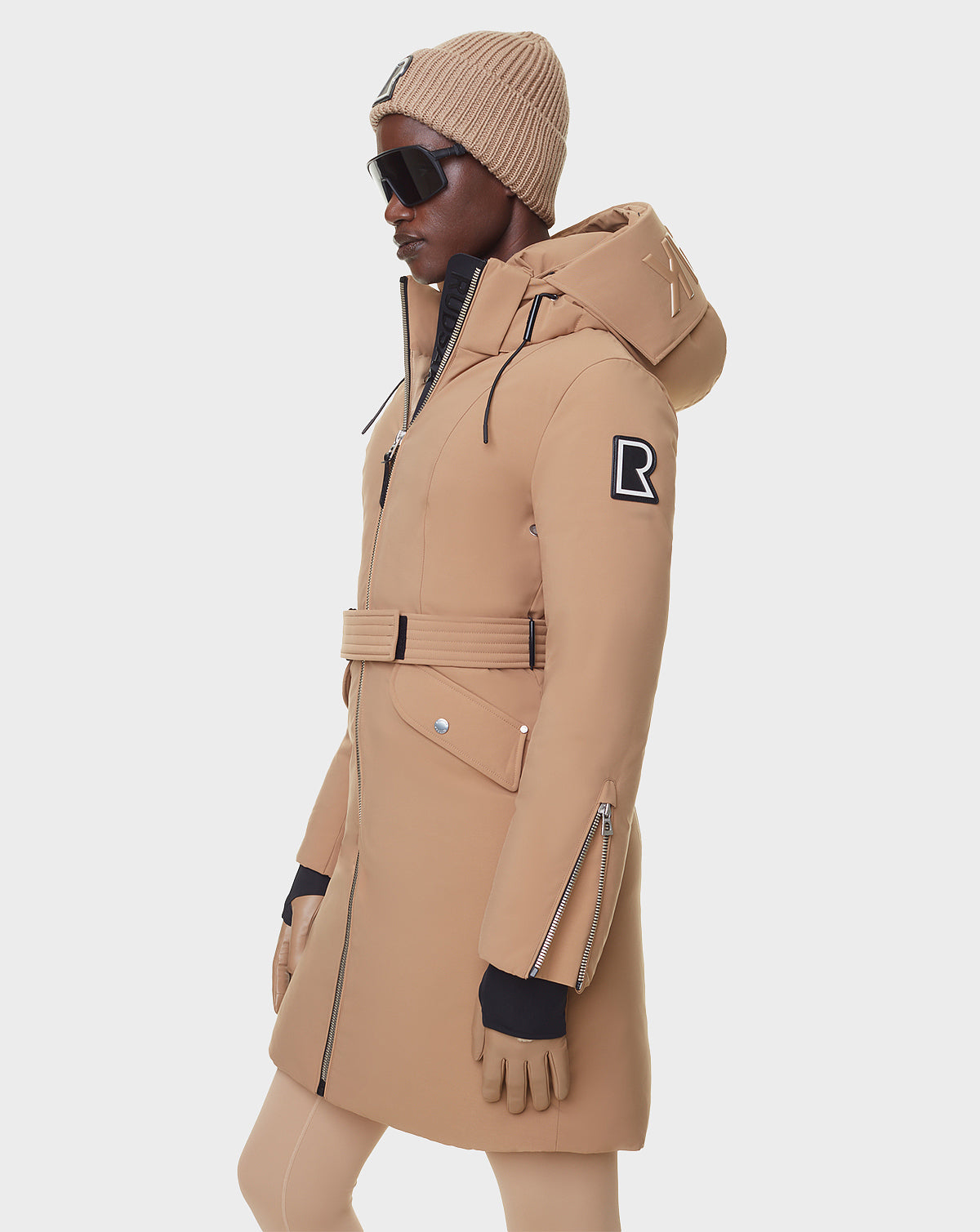 A woman in profile wears the RUDSAK MILENA - 8122536 TOFFEE winter coat with a belt, hood, matching knit beanie, black sunglasses and gloves. The toffee coat features an “R” patch on the upper arm and is shown against a plain white background.
