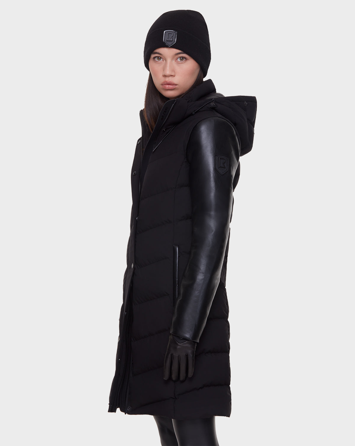 A woman in the Rudsak ELSA - L121519 BLACK sustainable long winter coat with leather sleeves, paired with black gloves and a black beanie, stands against a plain light background, looking toward the camera.
