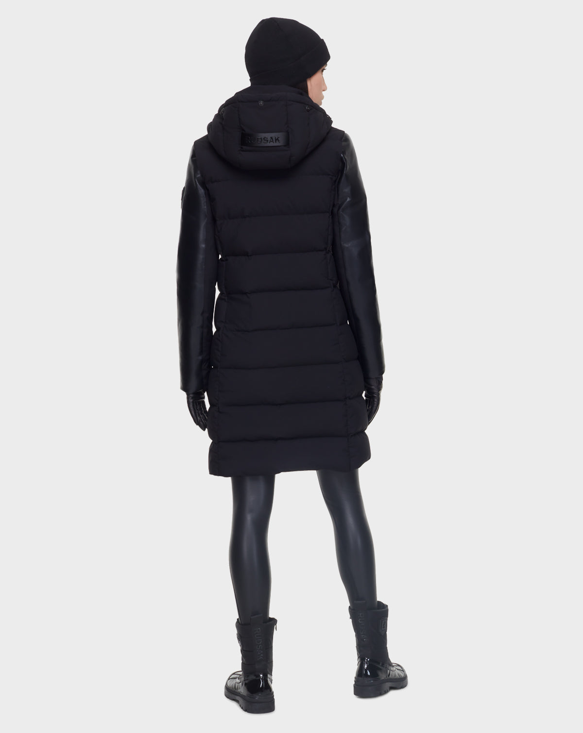 A person wearing the Rudsak ELSA - L121519 BLACK women’s quilted down puffer coat, along with black gloves, pants, and boots, stands facing away from the camera against a plain light background.