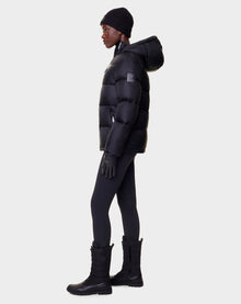 A person in profile stands against a plain light background wearing the Rudsak EVIE - L122517 BLACK womens down puffer jacket (RDS-certified) with a hood, black leggings, winter boots, gloves, and a beanie.