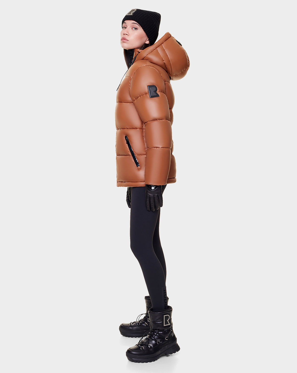 A woman in the Rudsak EVIE - L122517 DARK CAMEL down puffer, with waterproof zippers, black leggings, gloves, beanie, and boots stands in profile against a plain white background.
