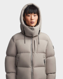 A person with straight black hair and bangs wears the Rudsak MAVE M - 8123572 Cement women’s down puffer jacket with hood up. The water-repellent jacket features zipped pockets and a high collar, standing against a light background.