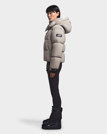 A person in profile wears the Rudsak MAVE M - 8123572 Cement women’s down puffer jacket with lightweight insulation, paired with black leggings and black winter boots, against a plain white background.