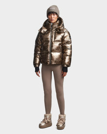 A person stands facing forward against a light background wearing the Rudsak MAVE S - 8123563 Metallic Ivory Camo womens down puffer jacket, taupe leggings, beige winter boots, and a light gray knit beanie.