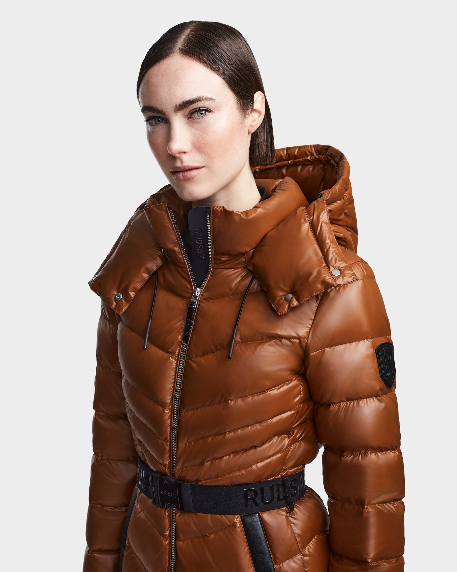 A woman wearing the Rudsak MALAYA - L123510 Dark Camel down puffer jacket with herringbone quilting and a black belt stands against a plain light gray background.