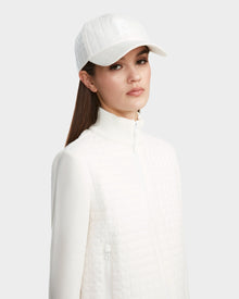 A woman wearing the Rudsak LOUISE - 8124046 Eggshell quilted jacket and matching white baseball cap stands against a plain light gray background, looking towards the camera with a neutral expression.