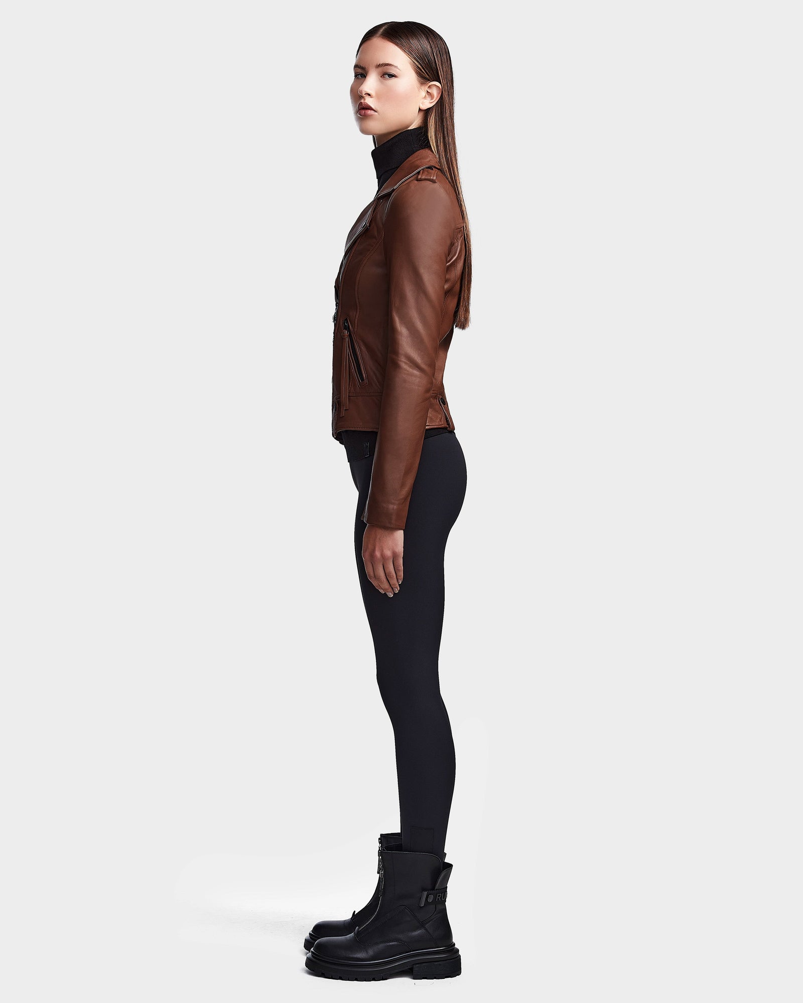 A woman stands in profile against a light gray background, wearing the RUDSAK MERGO - 8122009 COHIBA lambskin leather jacket, a black turtleneck, black leggings, and boots. Her straight hair is loose and her arms rest at her sides.