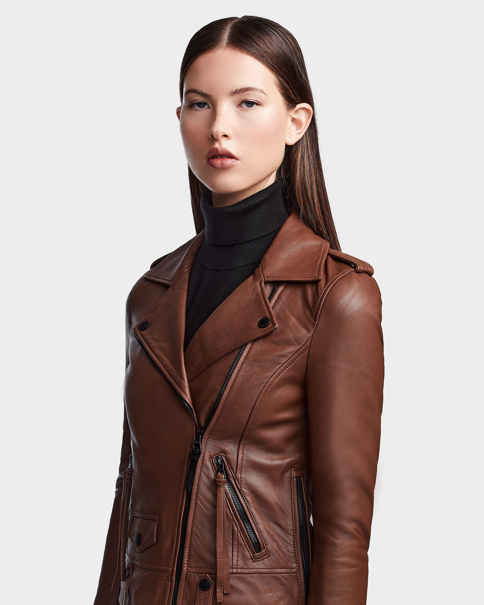 A young woman with straight brown hair wears the RUDSAK MERGO - 8122009 COHIBA lambskin leather jacket over a black turtleneck, posing confidently against a plain white background.