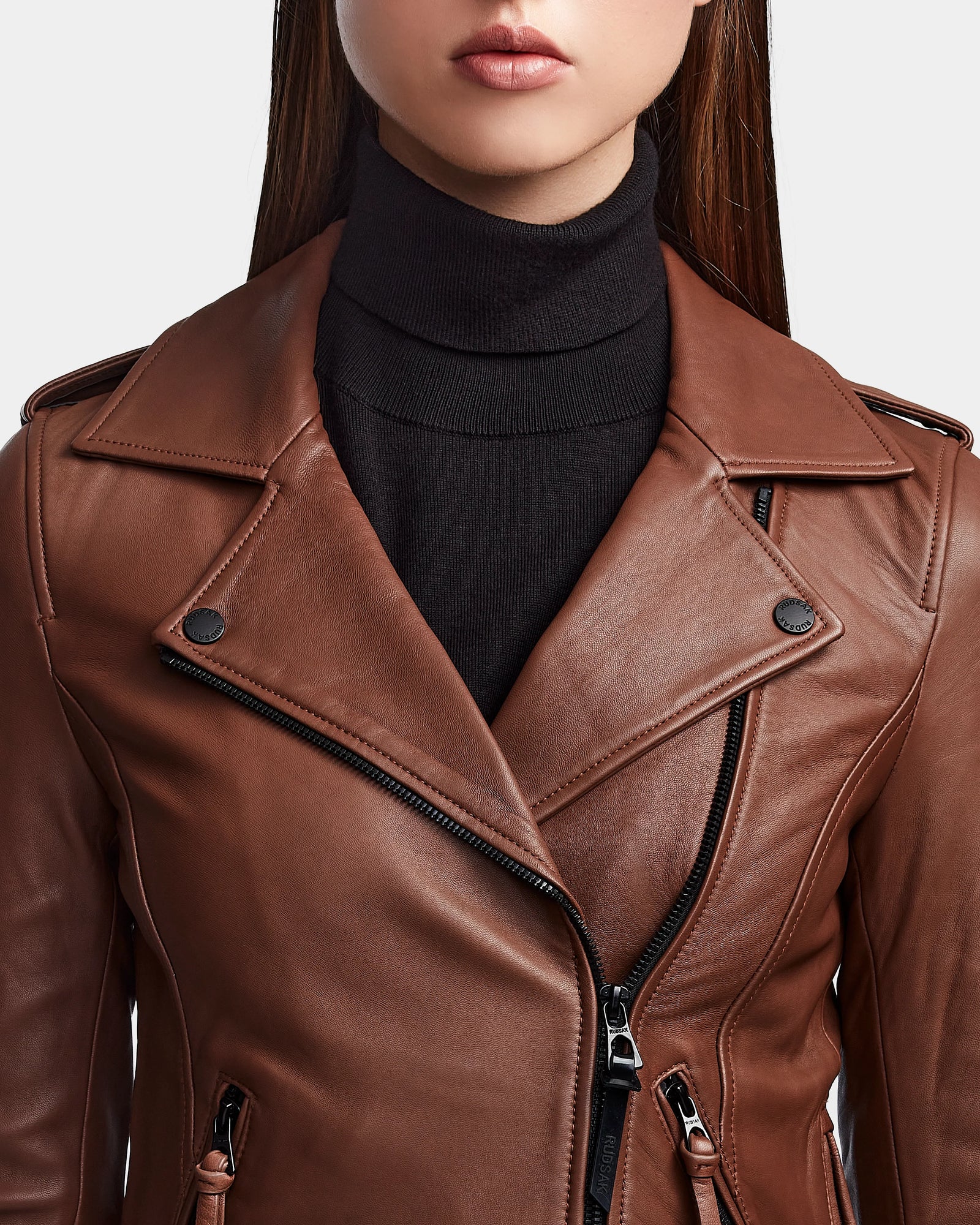 A woman wears the RUDSAK MERGO - 8122009 COHIBA biker jacket made from lambskin leather over a black turtleneck. The jacket has a diagonal zipper and button accents. Her lips and straight brown hair are partially visible.