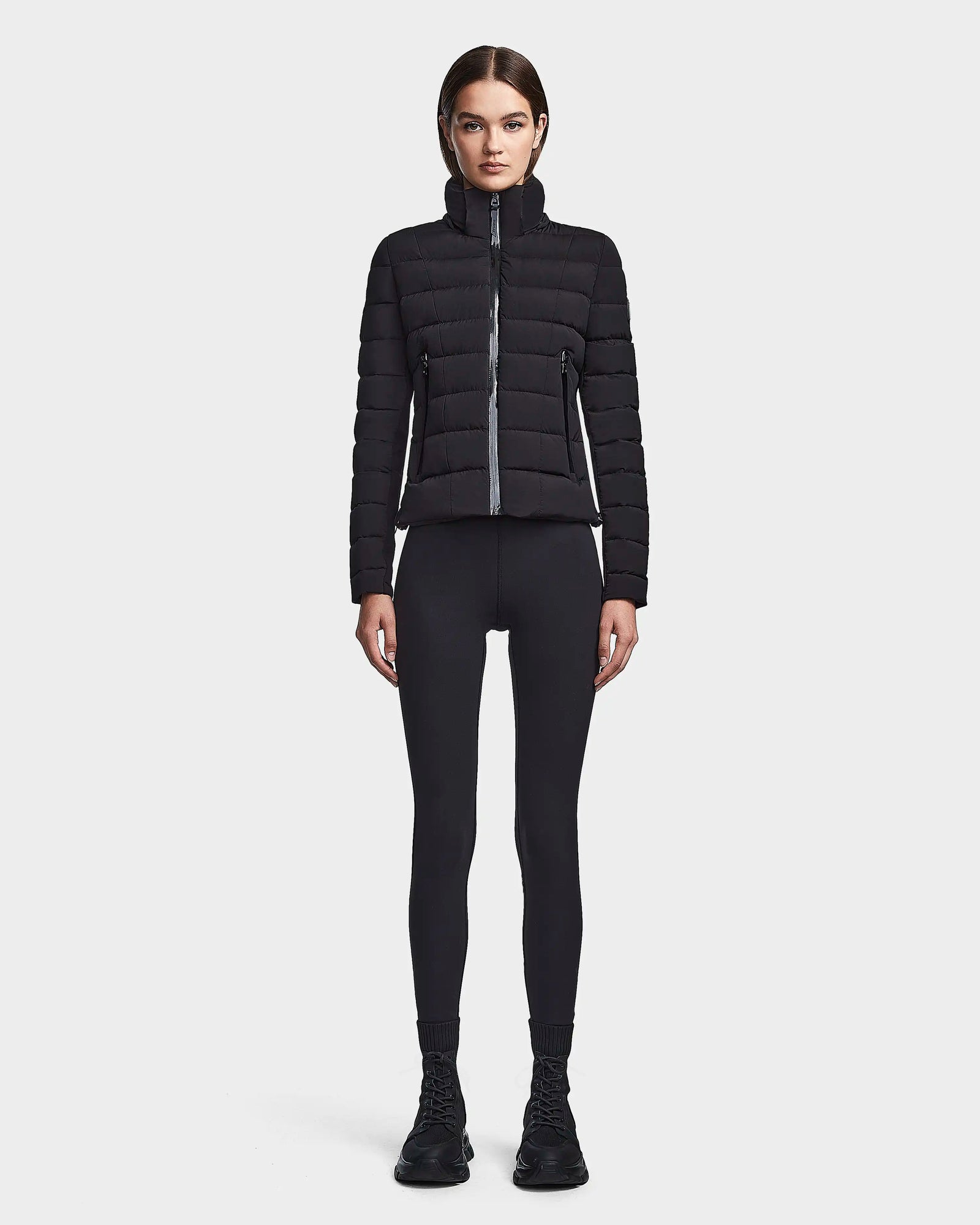 A woman faces forward against a plain white background, wearing the RUDSAK MELISSA - 8122551 BLACK puffer jacket with black leggings, socks, and sneakers. Her hair is pulled back and her arms rest at her sides.