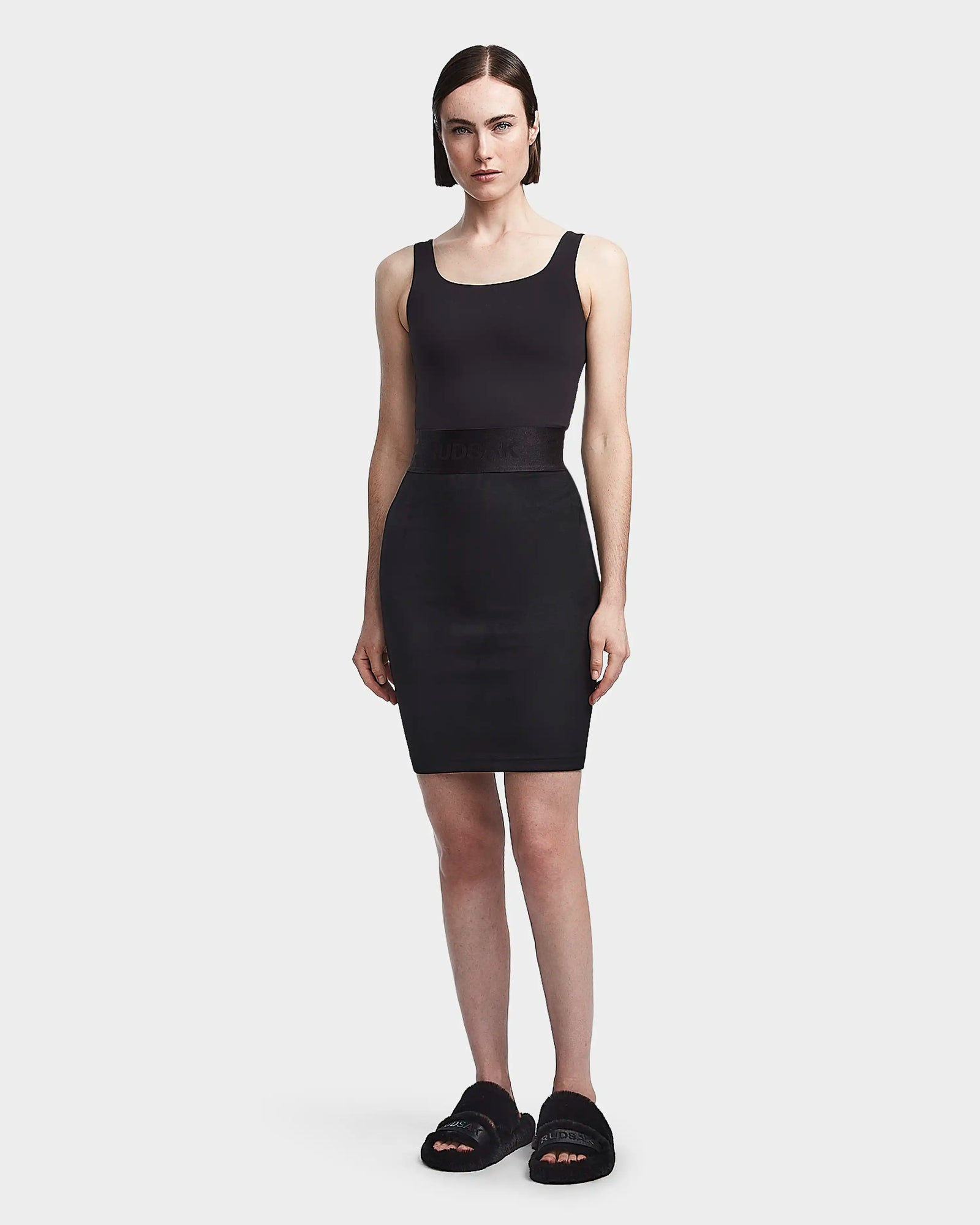A woman with straight dark hair faces forward in a sleeveless black tank top, the Rudsak CHEYNA - 8523514 Black womens pencil skirt, and black slide sandals, against a plain white background.