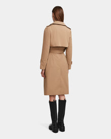 A woman with straight brown hair faces away, wearing the Rudsak KINDA - 8124690 Camel water-repellant trench and knee-high black boots against a plain white background.