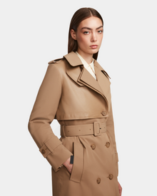 A woman with straight brown hair models the Rudsak KINDA - 8124690 Camel trench coat over a white collared shirt, standing against a plain white background and gazing slightly to the side.