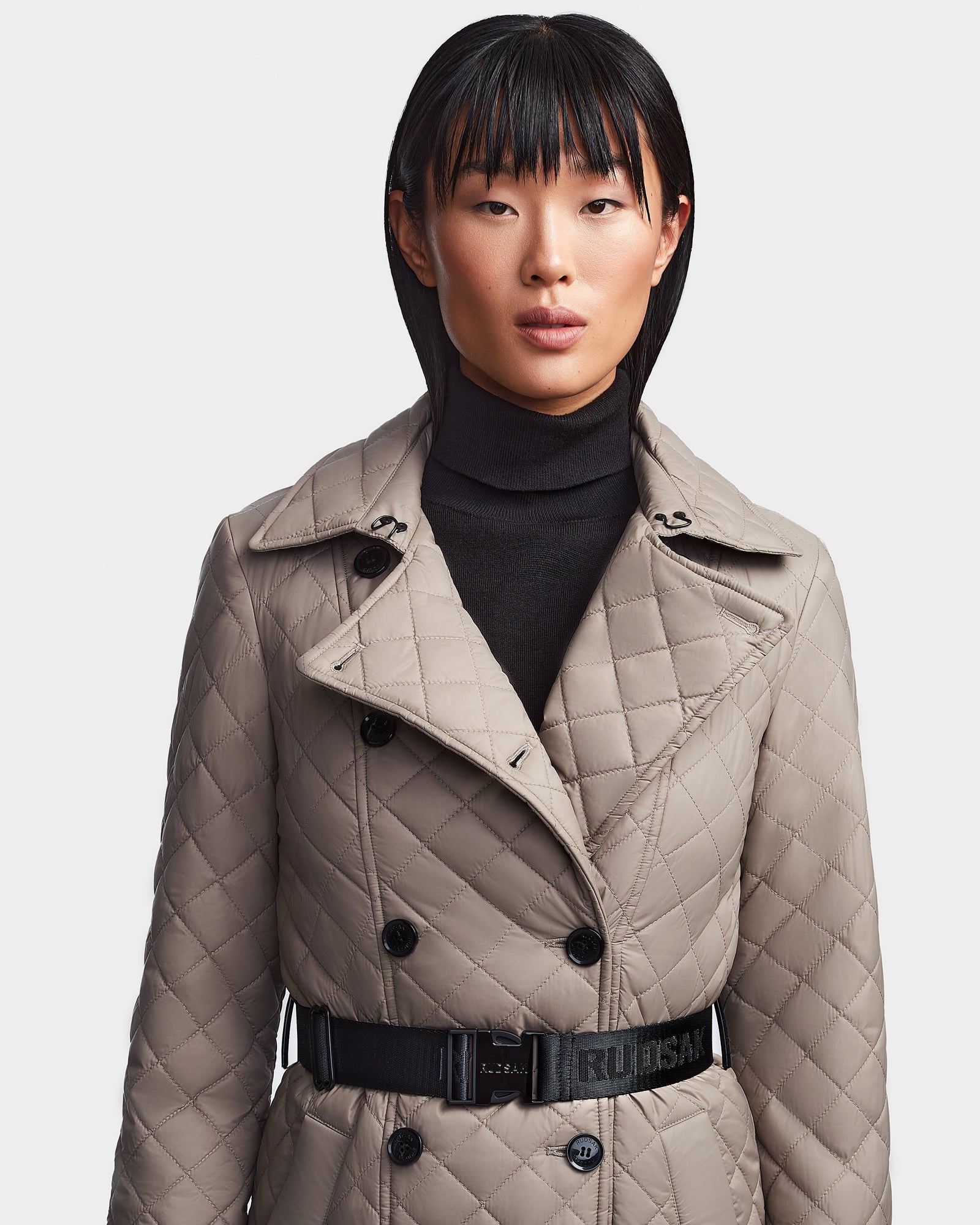 A person with straight, dark hair and blunt bangs wears the Rudsak DAISY - 8123580 Cement women's quilted maxi trench coat with a belted waist over a black turtleneck, standing against a plain light background.