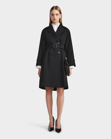 A woman with straight, shoulder-length hair and a neutral expression faces forward, wearing the Rudsak MIRELLE - 8124055 Black trench over a white shirt, black pointed heels, and holding a small black handbag.