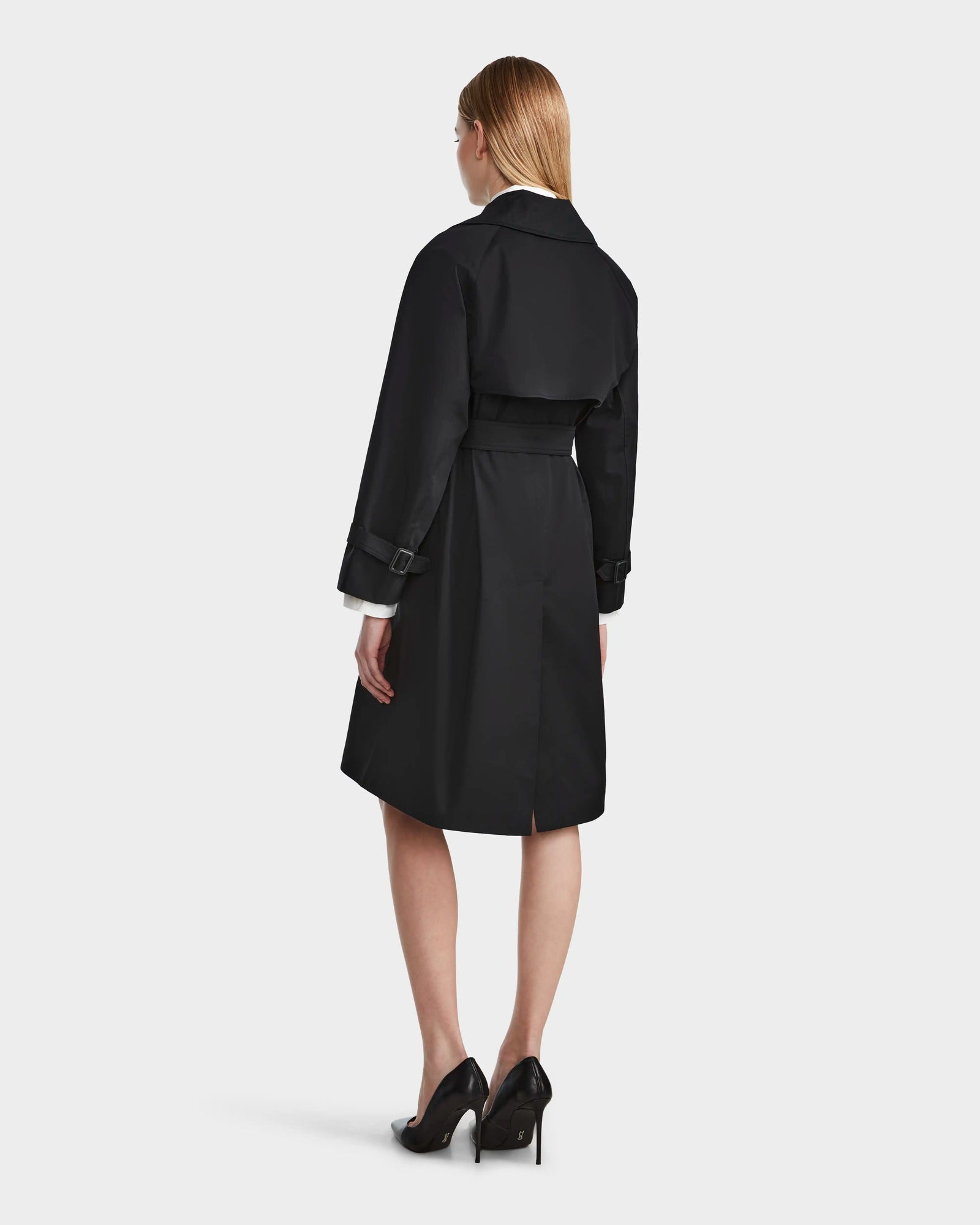 A woman with straight, light brown hair models the Rudsak MIRELLE - 8124055 Black women’s classic trench with a belted waist and black high heels, posing with her back to the camera against a plain light background.