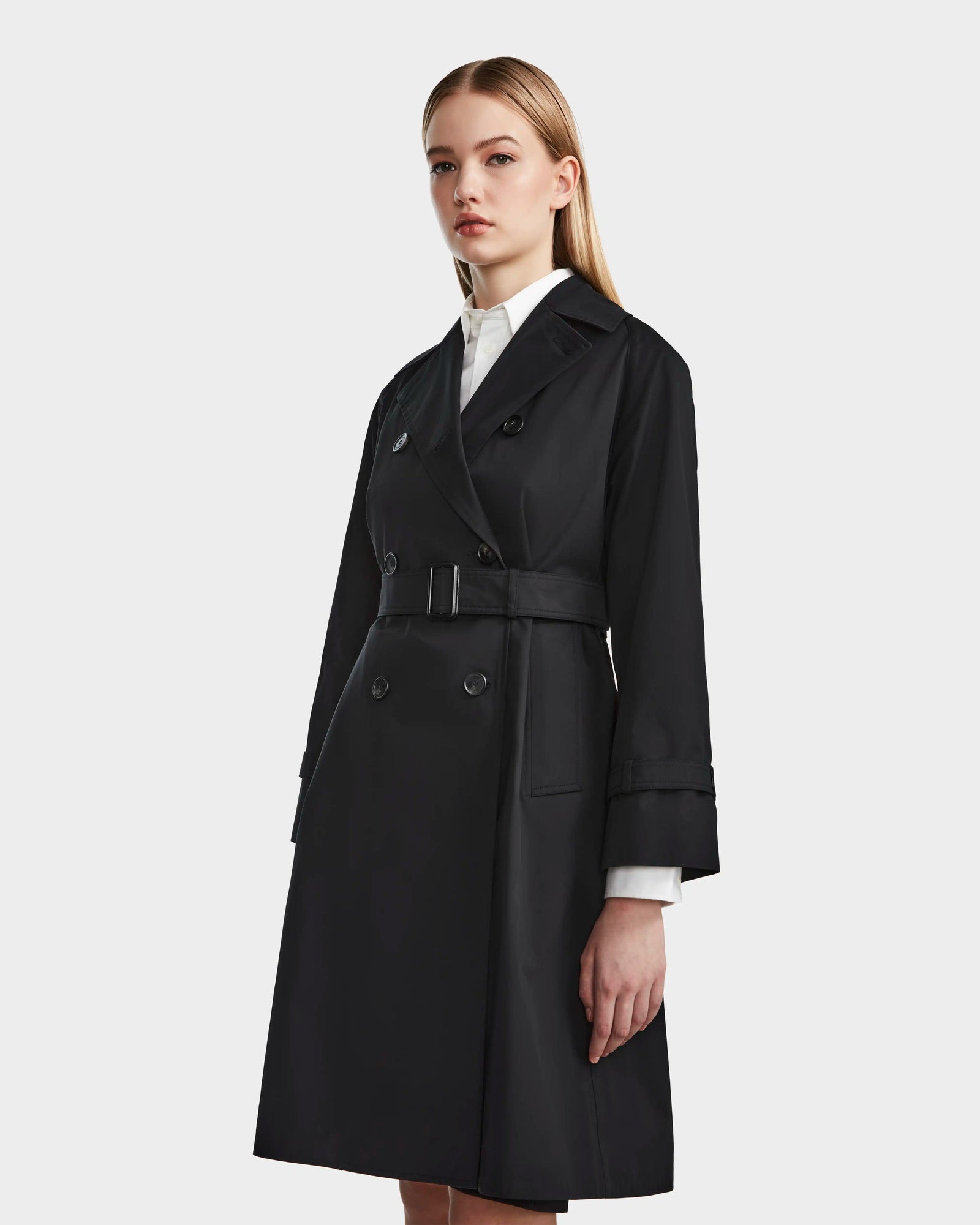 A woman with straight, long hair and a neutral expression stands against a light background wearing the Rudsak MIRELLE - 8124055 Black womens classic trench coat with a tailored silhouette, belted waist, and white shirt cuffs for versatile styling.