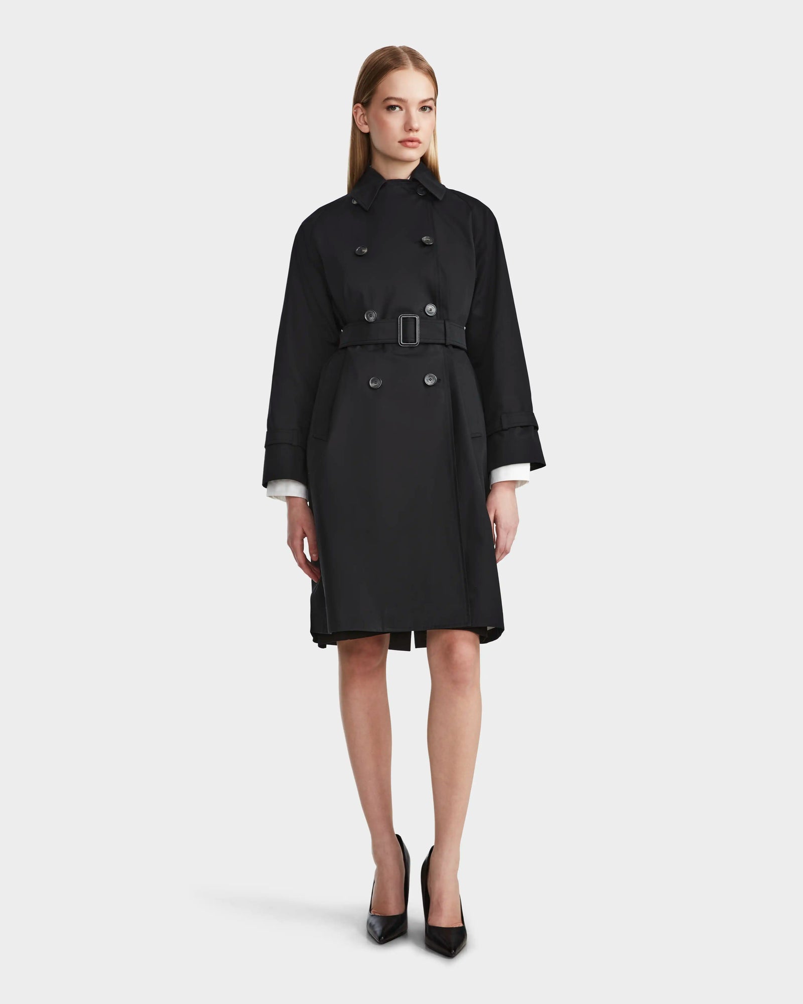 A woman faces forward in the Rudsak MIRELLE - 8124055 Black trench with a tailored silhouette and black heels, wearing a neutral expression against a plain white background.