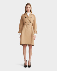 A woman stands facing forward in the Rudsak MIRELLE - 8124055 Khaki trench coat, knee-length with a tailored silhouette and belt, white shirt cuffs showing, paired with black high heels against a plain light background.
