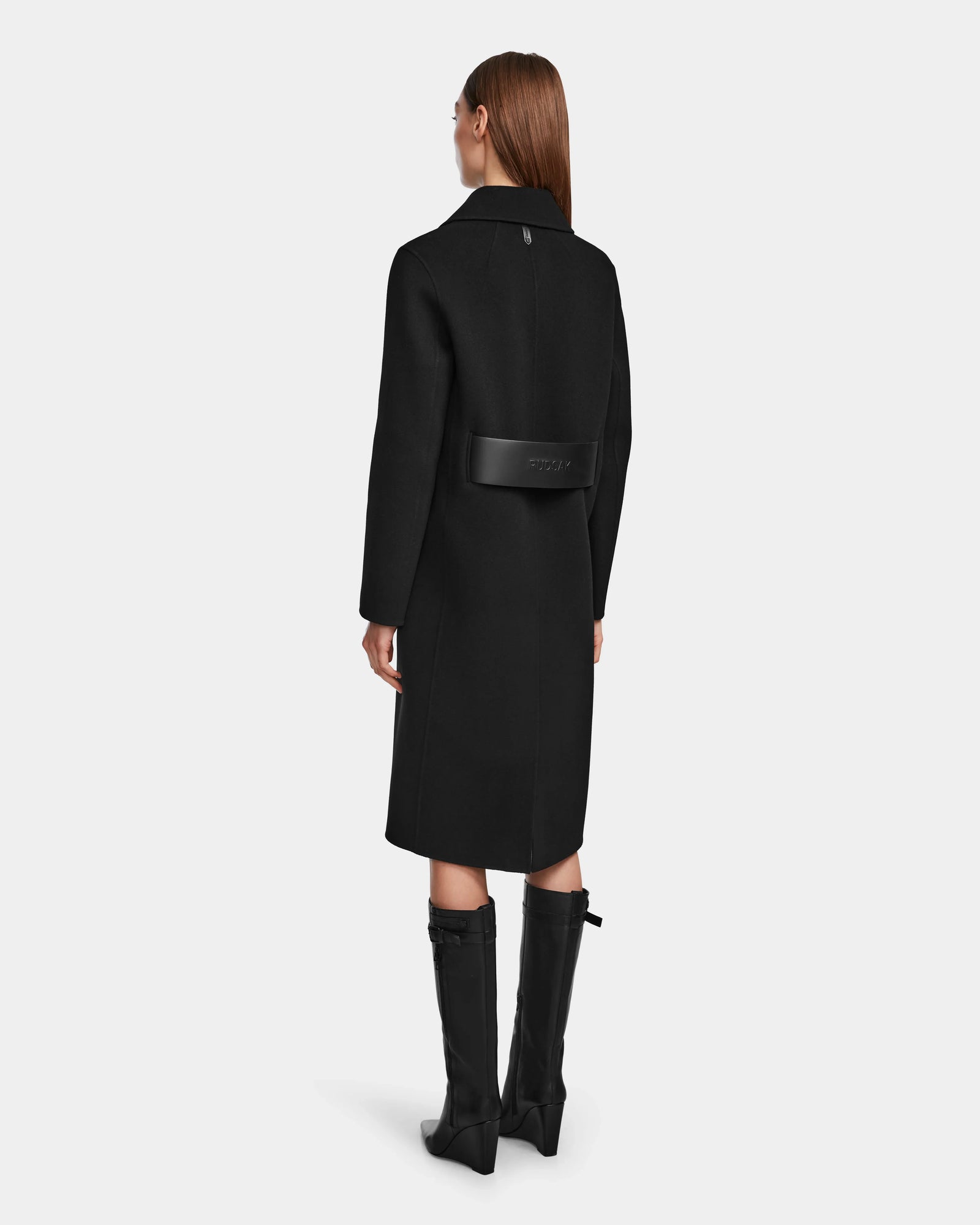 A woman with straight brown hair models the Rudsak LAYLAH - 8124699 Black knee-length women’s wool coat featuring a wide back belt and wears knee-high black boots, standing with her back to a plain light gray background.