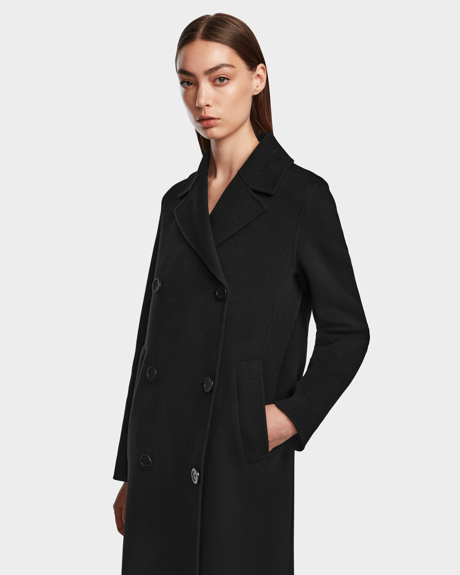 A woman with straight brown hair and a neutral expression stands against a light background wearing the Rudsak LAYLAH - 8124699 Black knee-length double-breasted coat with a classic collar, one hand tucked in her pocket.