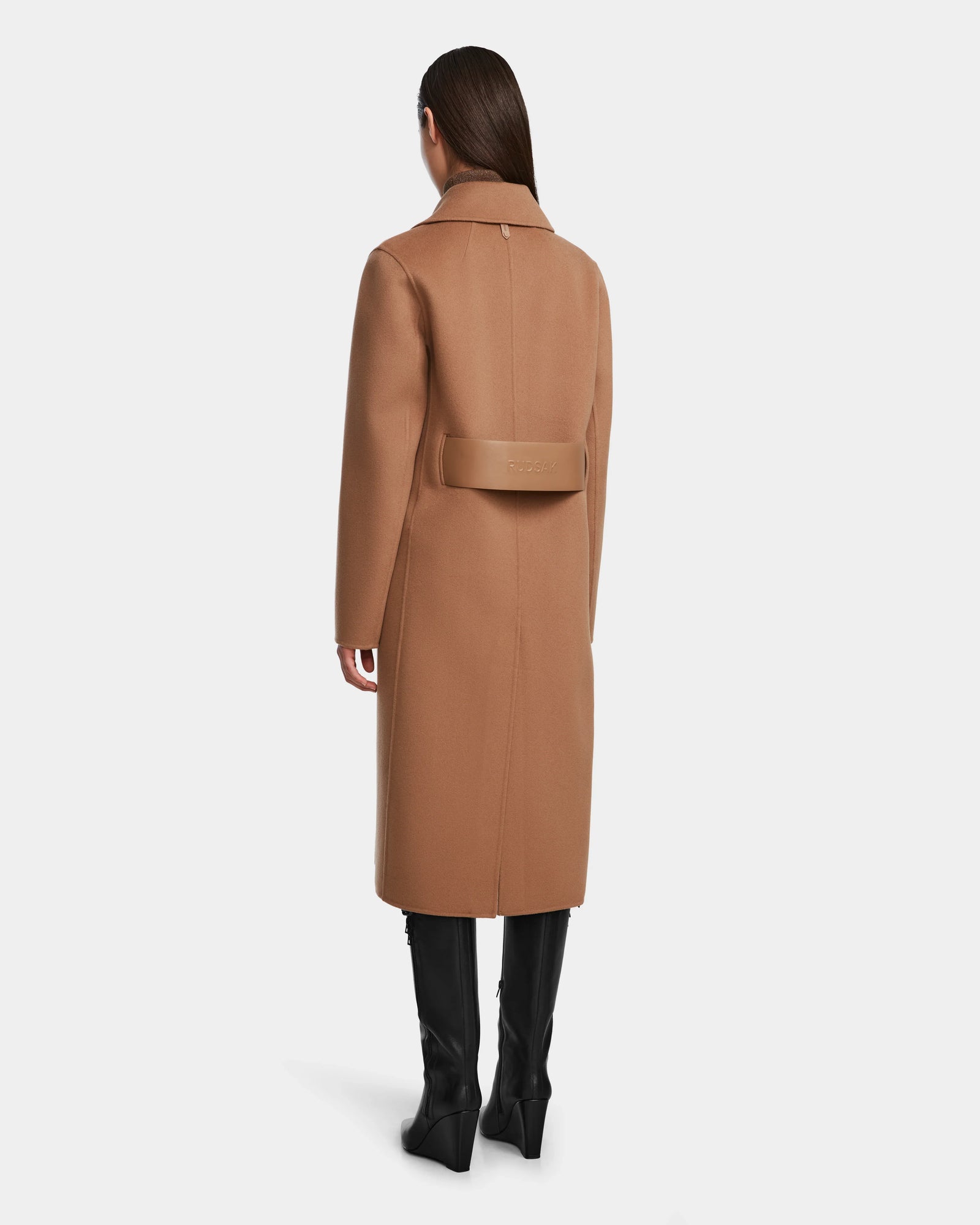 A woman faces away, wearing the Rudsak LAYLAH - 8124699 Camel knee-length wool coat with a wide back belt and black knee-high boots. Her straight hair is down, set against a plain white background.