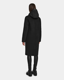 A woman faces away, wearing the Rudsak ARLA - 8124579 black womens wool coat with leather sleeves, paired with black leggings and boots, against a light gray background.