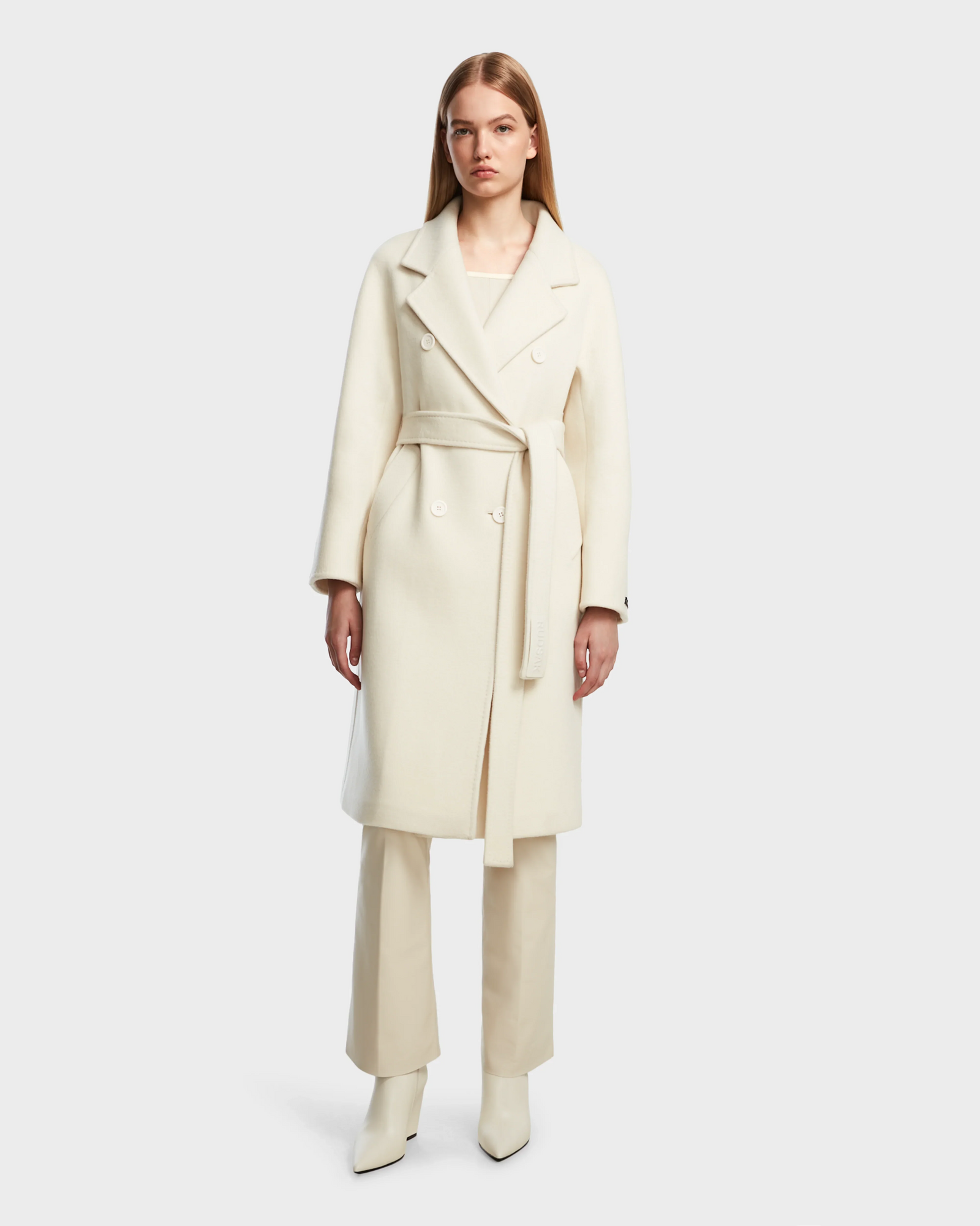 A woman with straight blonde hair models the Rudsak KOURT W - 8124702 Iceland long, double-breasted belted wool coat over cream pants and matching heeled boots against a plain light gray background.