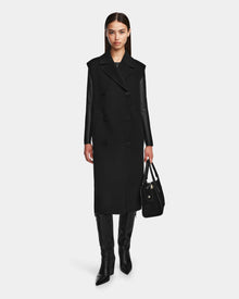 A woman models the Rudsak NOURA - 8124698 Black, a sleek, knee-length double-breasted vest, styled over a black top with knee-high boots and a black handbag against a plain white background.