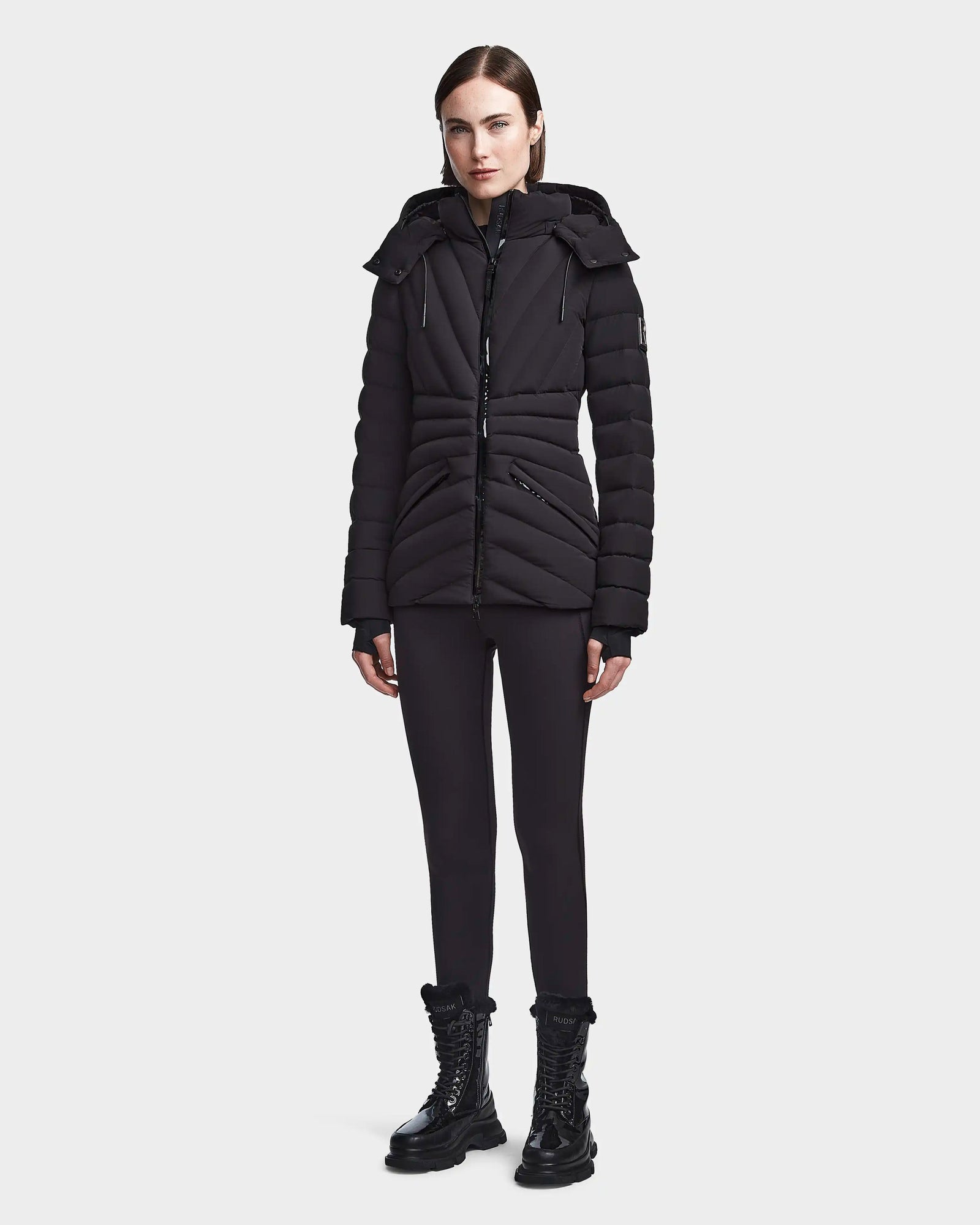 A woman faces forward in the Rudsak HANNA - 8123562 Black fitted winter down jacket with hood, made from SMART STRETCH fabric, styled with black leggings and lace-up boots against a plain light background.