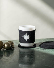 A white candle in a black star-cutout holder rests on a green marble surface, echoing Star Glas sleeves. Nearby are two gold spheres and stacked black coasters by graf lantz. Soft sunlight creates gentle shadows in the background.