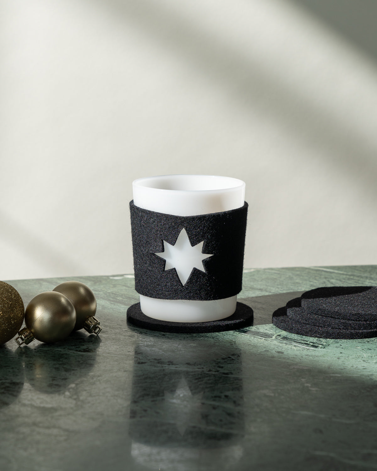 A white candle in a black star-cutout holder rests on a green marble surface, echoing Star Glas sleeves. Nearby are two gold spheres and stacked black coasters by graf lantz. Soft sunlight creates gentle shadows in the background.
