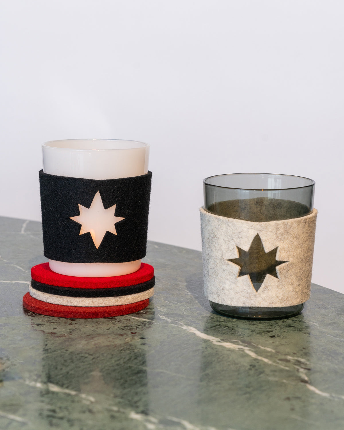 Two glasses in felt Star Glas sleeves with star cutouts rest on a marble surface. Made from Merino wool, one sleeve is black and the other is white. Three coasters in black, red, and white are stacked beside them. The graf lantz set is Oeko-Tex Certified for sustainability.
