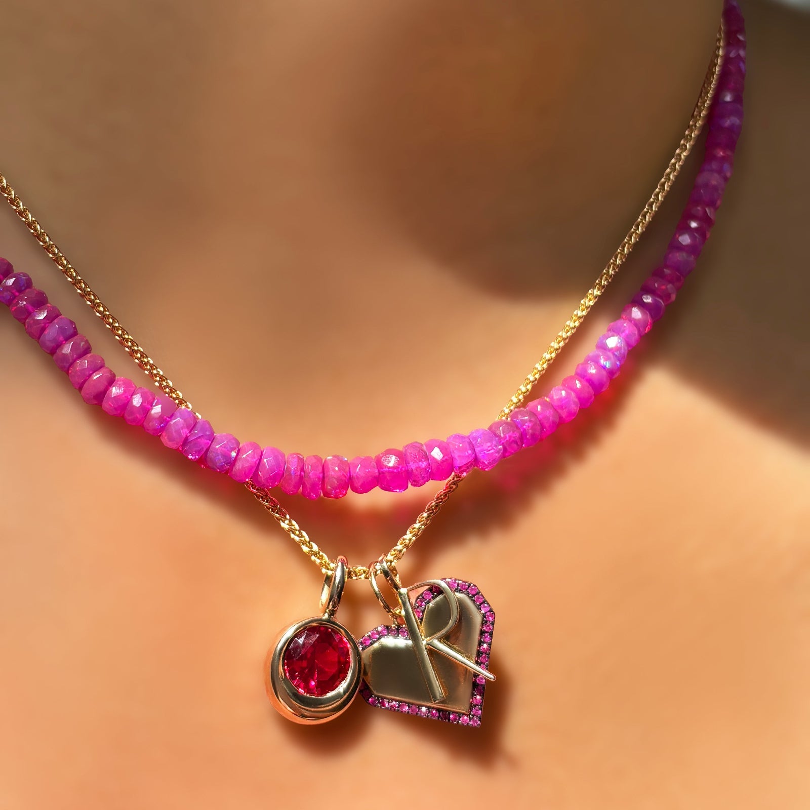 Fuchsia faceted opal necklace layered over a wheat chain with molten solitaire charm with ruby, digi heart charm with ruby border, and quill letter R charm on the neck.
