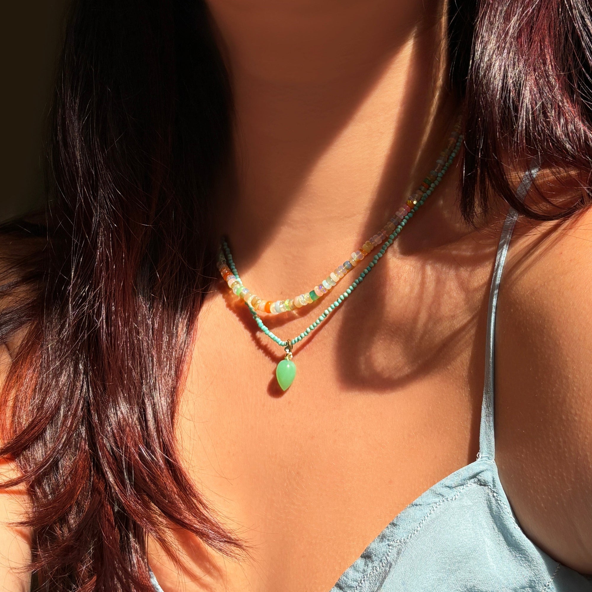 Shimmering beaded necklace made of smooth opal rondels in shades of tan, teal, orange and pink on a gold linking ovals clasp. Styled on a neck layered with a turquoise slim gemstone and a acorn drop charm.