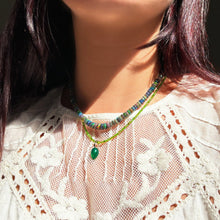 Shimmering beaded necklace made of faceted opals in shades of green, yellow, and blue on a gold linking ovals clasp, layered with a gemstone necklace and acorn, on a neck.