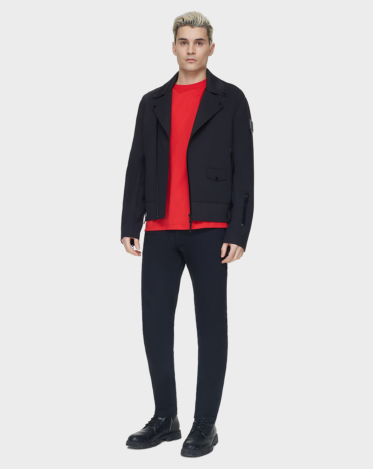 A young man with short blonde hair models the RUDSAK JUNE - 6122005 BLACK mens jacket over a bright red shirt, black pants, and black shoes, standing against a plain white background.