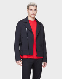 A man with short, light blond hair models the RUDSAK JUNE - 6122005 BLACK biker jacket over a bright red shirt and black pants against a plain light gray background.