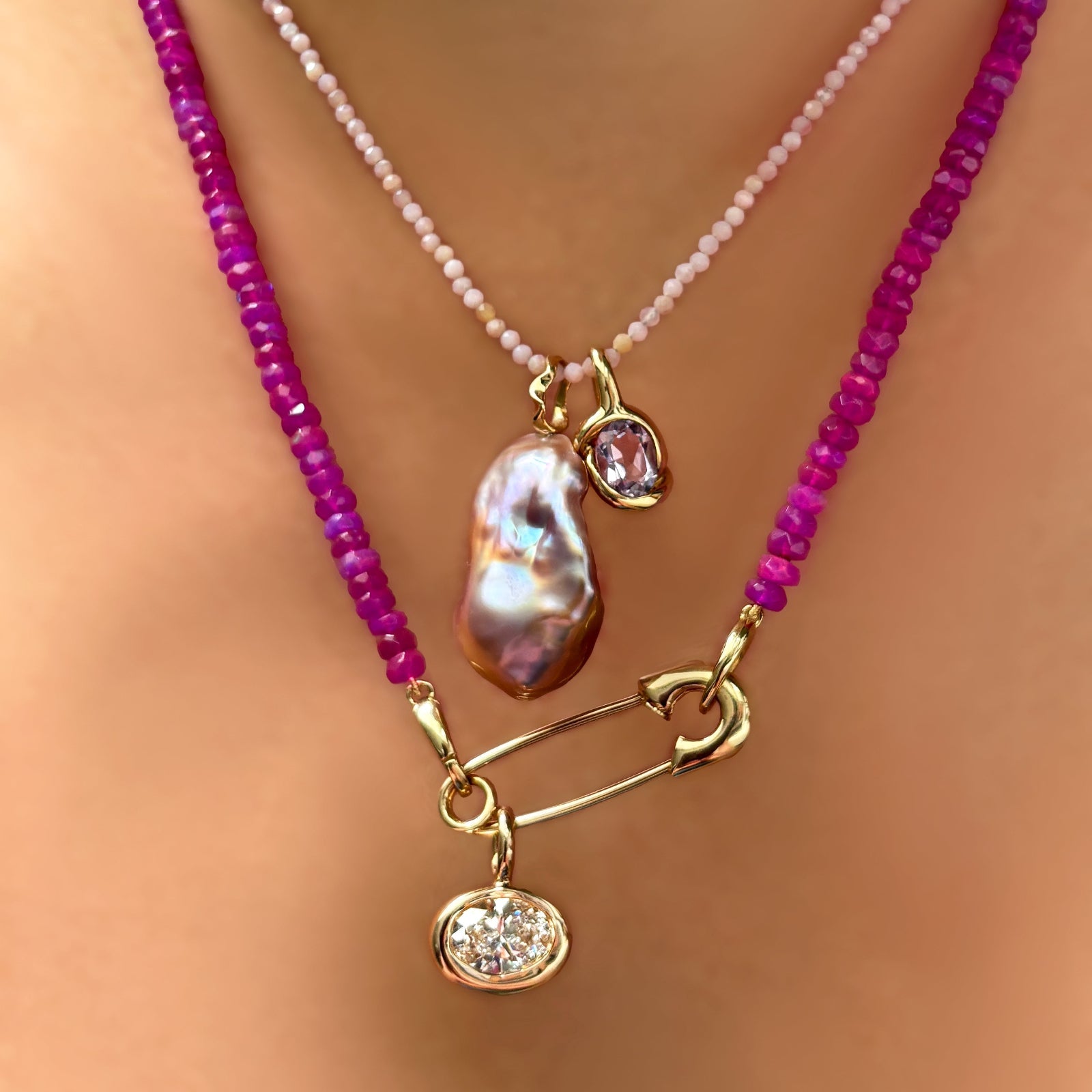 slim gemstone necklace in pink opal with an amethyst knot charm + blush baroque pearl charm, layered with a safety pin charm lock on a fuchsia faceted opal necklace with an oval diamond solitaire charm hanging from it.