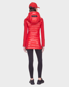 A person faces away, wearing the Rudsak MERINA - 8121006 POPPY RED women’s light jacket with a hood and matching red cap, paired with black leggings and sneakers against a plain light background.