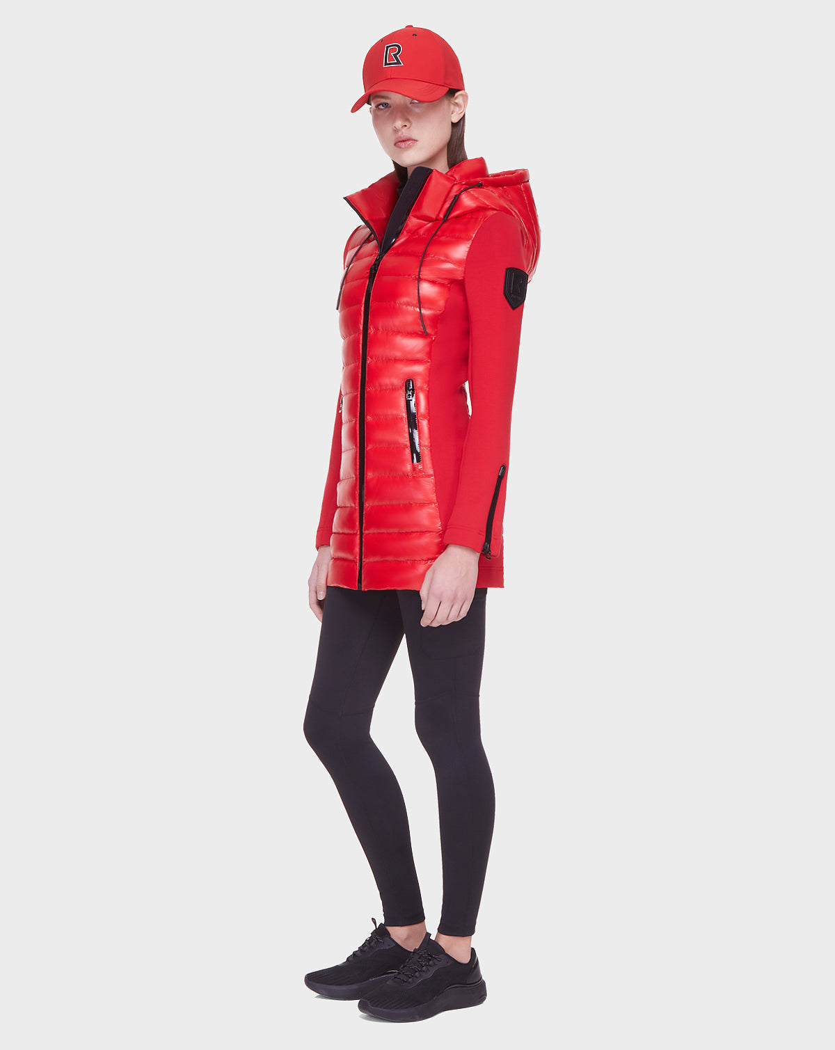 A woman wearing the Rudsak MERINA - 8121006 POPPY RED quilted down jacket stands against a plain white background, styled with a red baseball cap, black leggings, and black sneakers.