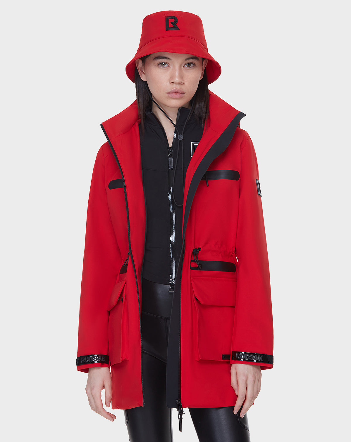 A woman stands against a plain white background wearing the Rudsak CAMY - 8122002 POPPY RED rain trench with black accents, a matching red bucket hat with an R logo, a black zip-up jacket underneath, and black pants.
