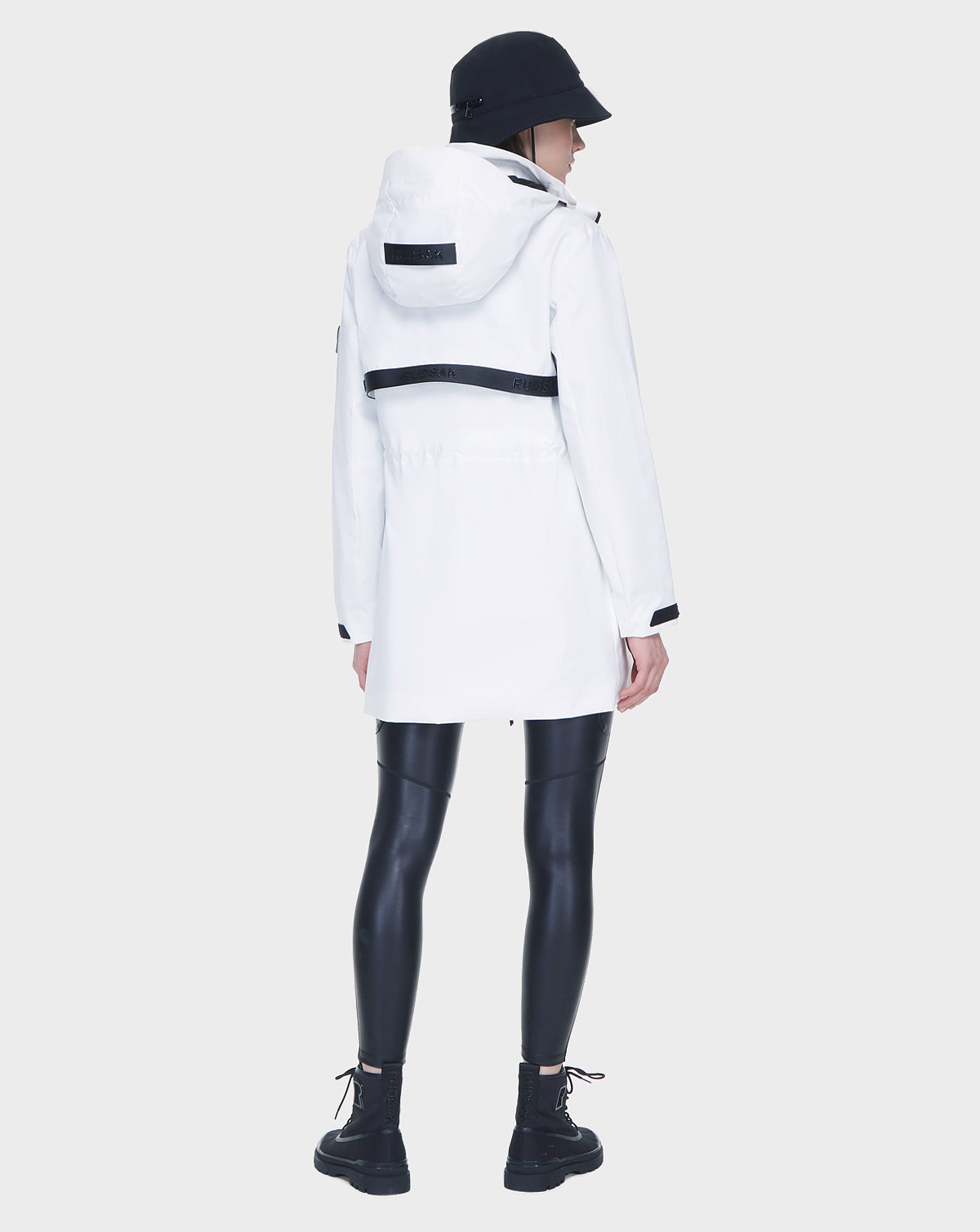 A person in the Rudsak CAMY - 8122002 WHITE water-resistant trench coat, black bucket hat, shiny black leggings, and black boots stands with their back to the camera against a plain light gray background.