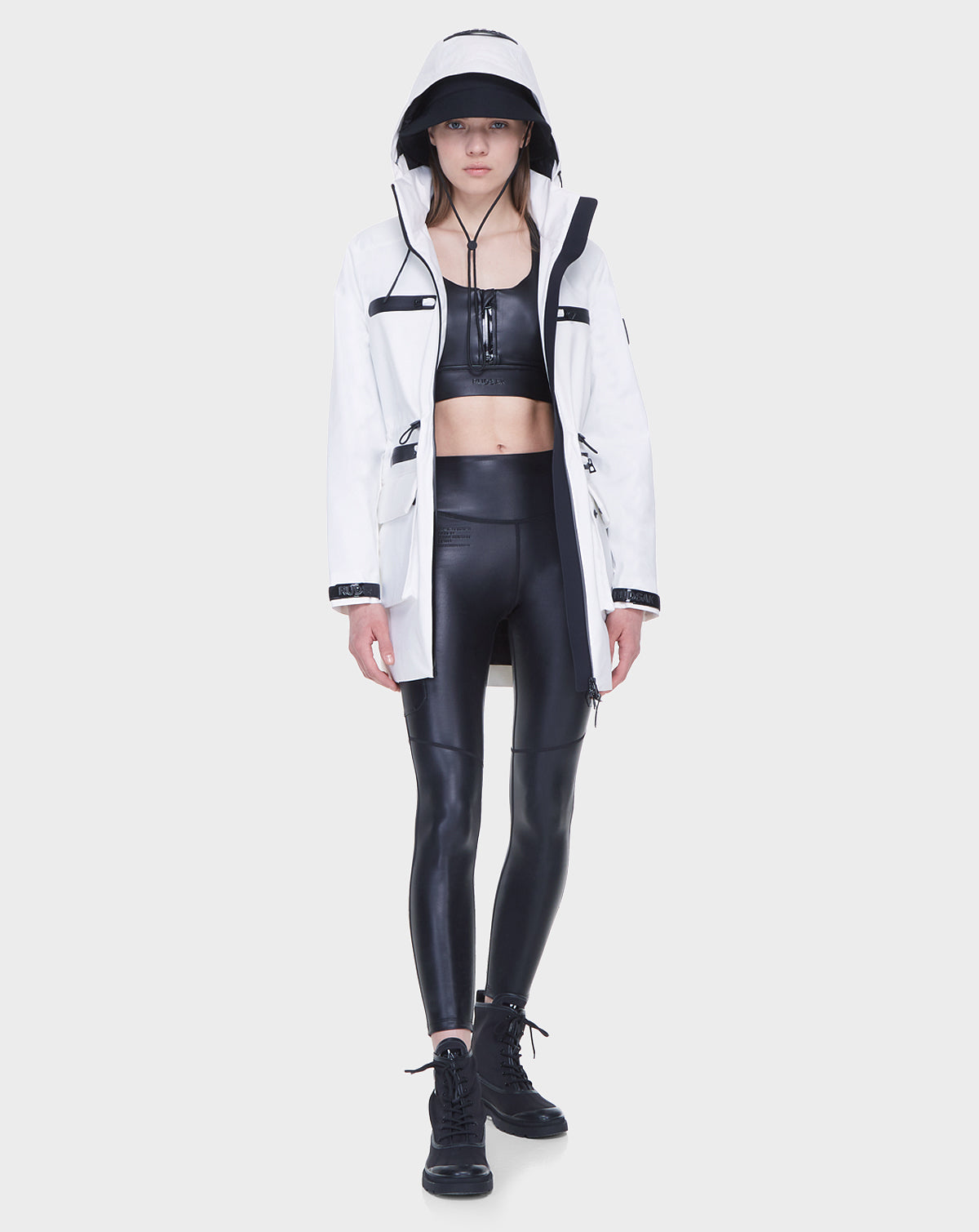 A woman faces forward wearing the Rudsak CAMY - 8122002 WHITE water-resistant trench coat over a black crop top, shiny black leggings, and black boots. The background is plain white.