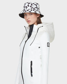 A woman wears the Rudsak CAMIRA - 8122011 WHITE womens activewear jacket with black zipper details and a black-and-white geometric bucket hat, standing against a plain light gray background—stylish comfort for outdoor workouts.